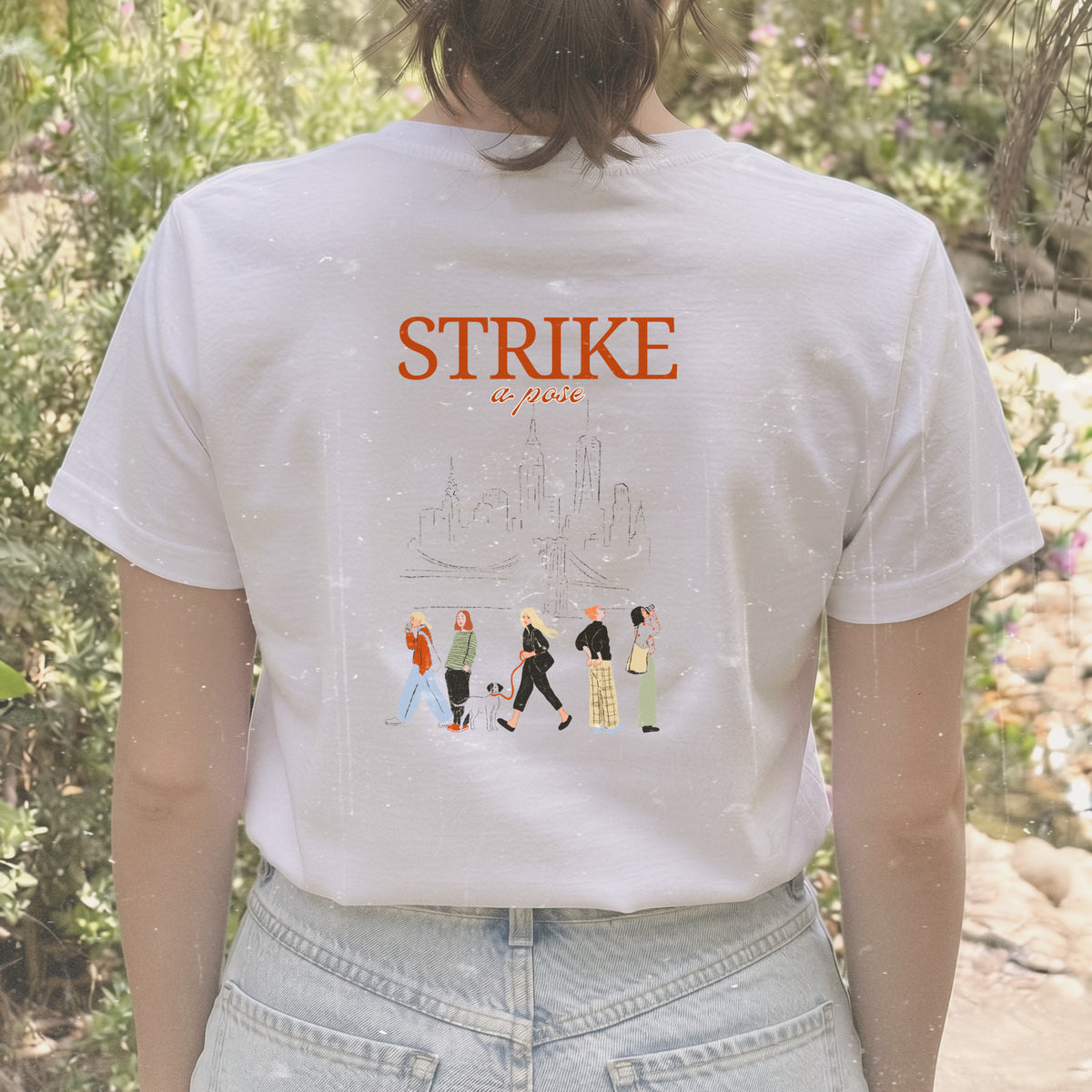 Camiseta de algodón orgánico de Laniñabowie modelo Carolyn en color blanco, con ilustración grande en la espalda con silueta de Nueva York, la frase "STRIKE A POSE" y mujeres caminando con referencia a Carolyn Bessette-Kennedy.